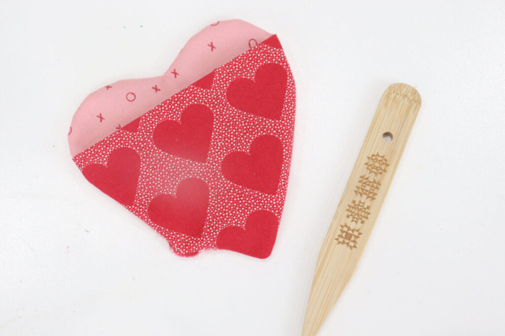 Image contains a pink fabric heart with a red fabric pocket. A wooden turning stick sits nearby on a white background.