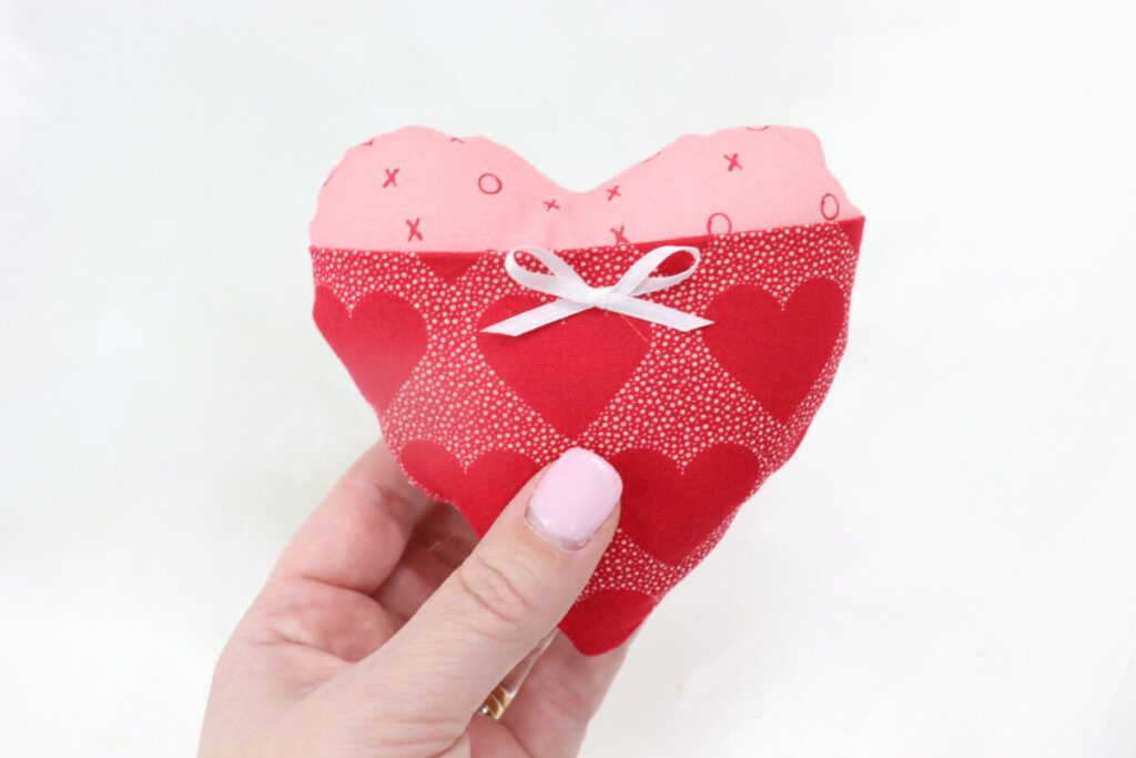 Image contains Amy’s hand holding a pink plush heart with a red fabric pocket and a white ribbon bow.