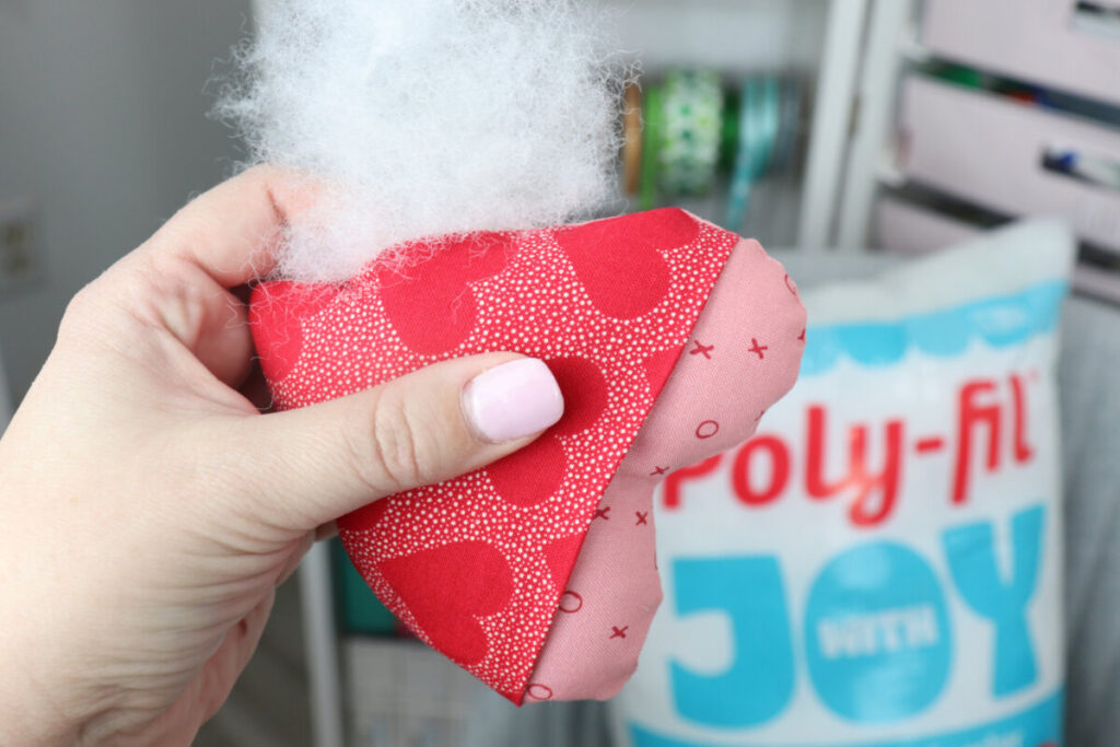 Image contains Amy’s hand adding Poly-Fil to the fabric pocket heart. A bag of Poly-Fil sits in the background.