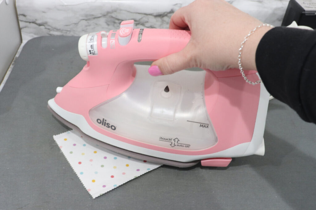 Image contains Amy’s hand holding a pink Oliso iron, ironing a piece of white polka dot fabric to fusible adhesive.