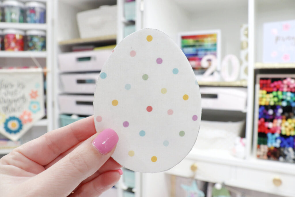 Image contains Amy’s hand holding a piece of white polka dot fabric cut into an egg shape in front of her DreamBox filled with assorted craft supplies.