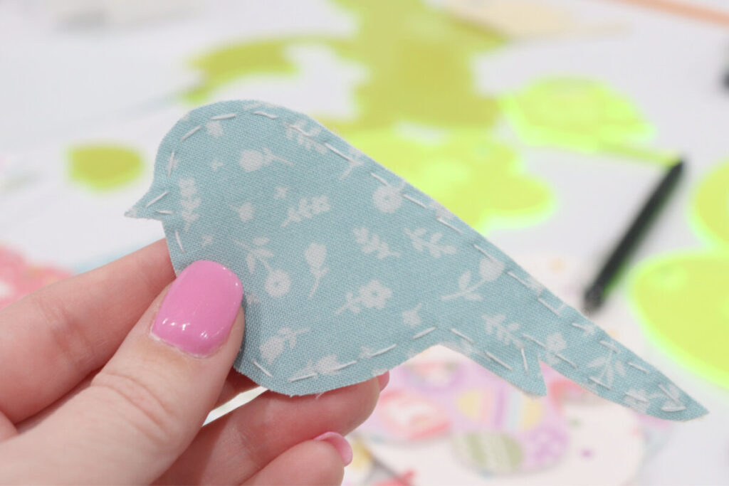 Image contains Amy’s hand holding a blue bird cut from fabric and hand stitched around the outer edge.