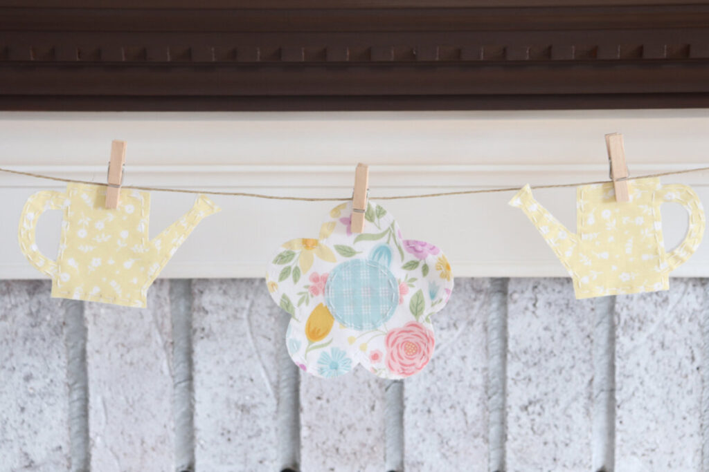 Image contains a closeup of the spring banner, showing the flower in the center and a yellow watering can on each side, clipped to twine with tiny clothespins.