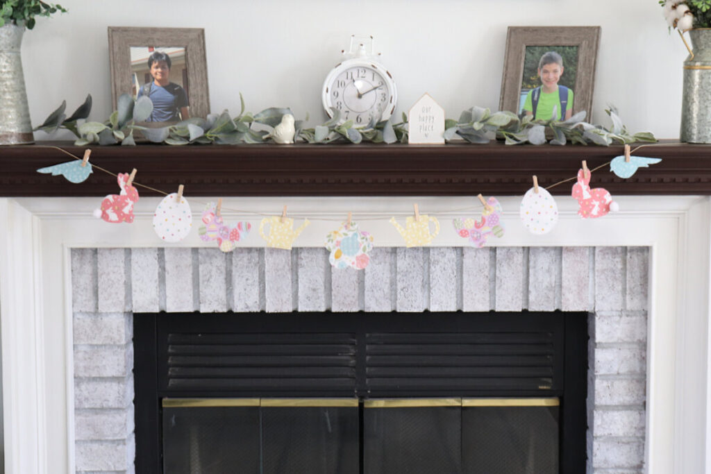 Image contains a white fireplace with a brown mantel, decorated with a spring banner made from multicolored fabric shapes.