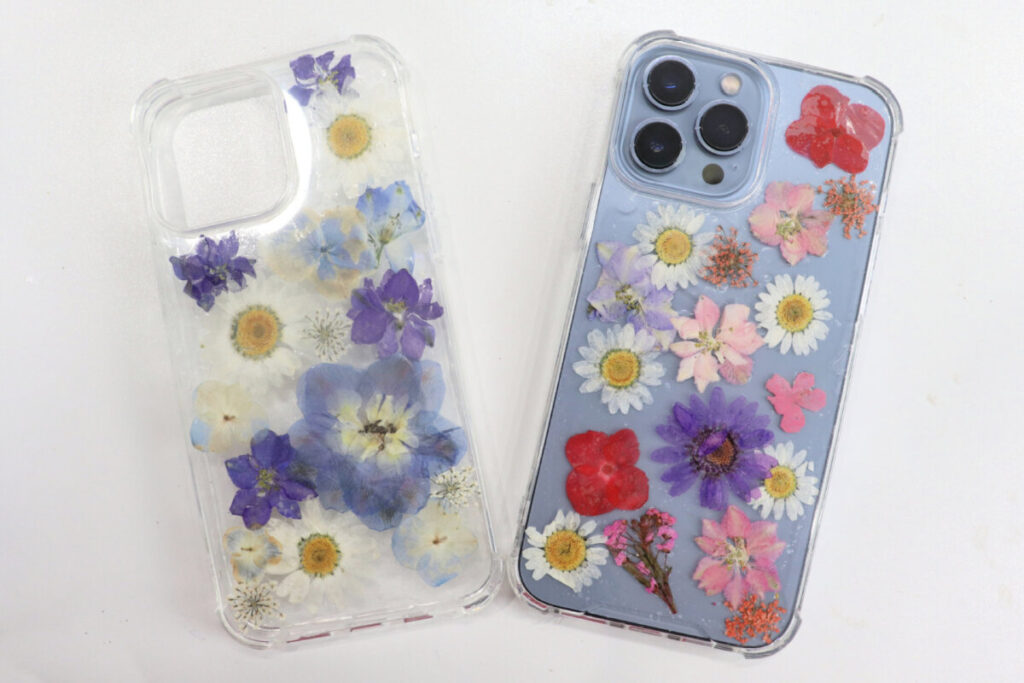 Image contains two clear phone cases decorated with pressed flowers ; one empty and one with a blue iPhone inside.
