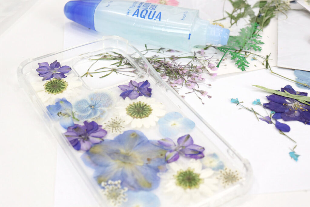 Image contains a clear phone case decorated with a variety of white, purple, and blue pressed flowers. More pressed flowers and a tube of glue sit off to the side on a white background.