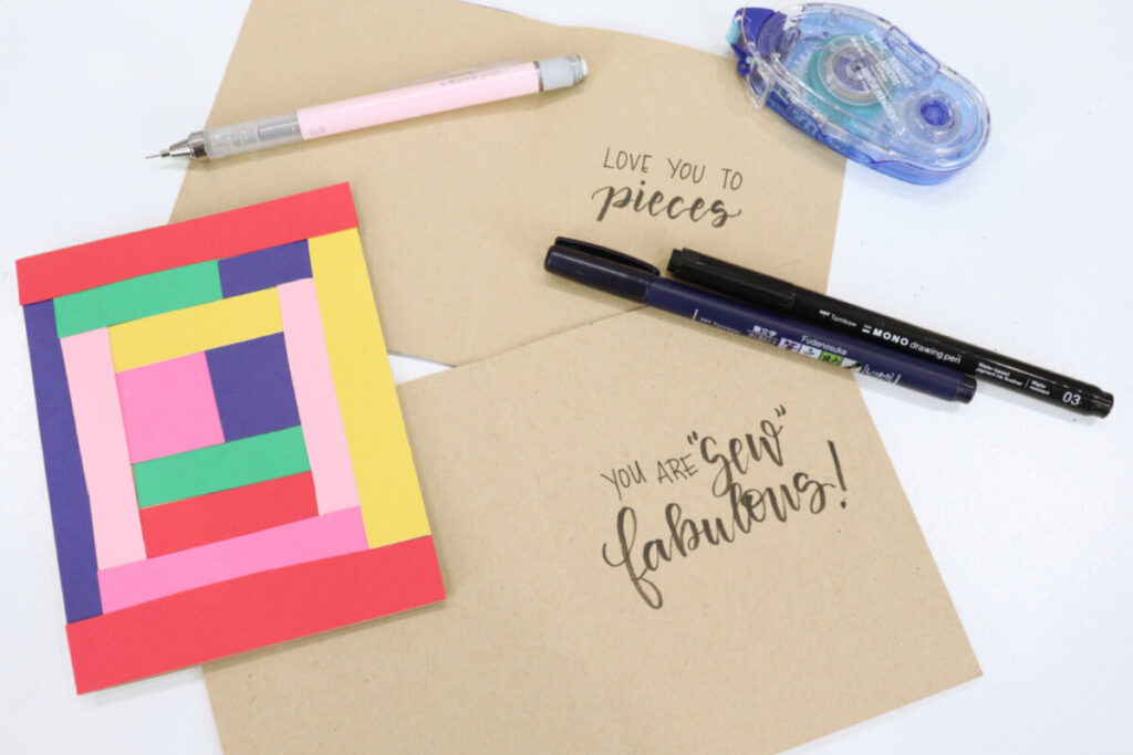 Image contains two kraft-colored cards opened to show the inside, where the phrases “love you to pieces” and “you are sew fabulous” are written in black ink. A closed card, decorated with colorful strips of cardstock in a geometric pattern, sits on top, along with a mechanical pencil, two black markers, and an adhesive runner.