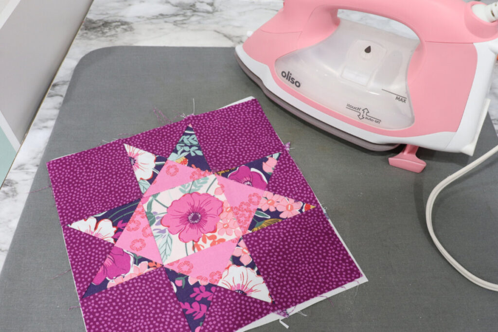 Image contains an Ohio Star quilt block made from purple and pink patterned fabrics on a grey pad next to a light pink Oliso iron.