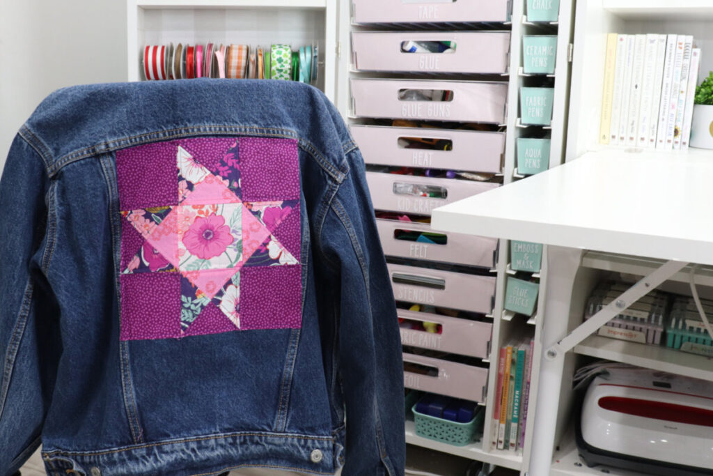 Image contains a denim jacket with a pink and purple quilt block on the back, draped over a chair in front of Amy’s DreamBox craft organization system.