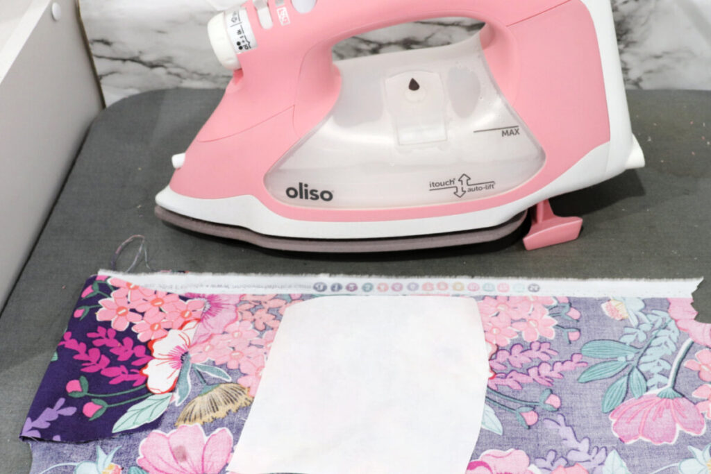 Image contains a pink Oliso iron on top of a grey ironing mat. A piece of floral fabric lays face down in front of it with a piece of fusible interfacing on top.