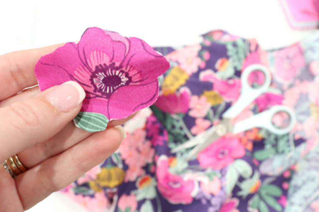 Image contains Amy’s hand holding a pink flower cut out from a piece of fabric. The piece of fabric and a pair of teal handled scissors are out of focus in the background on top of a white table.