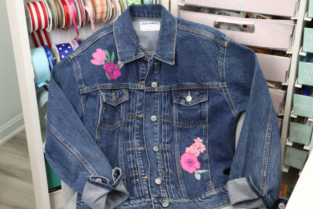 Image contains a denim jacket with bright pink and purple flowers added to the front as embellishments. It is draped over a grey chair in front of a cabinet full of craft supplies.