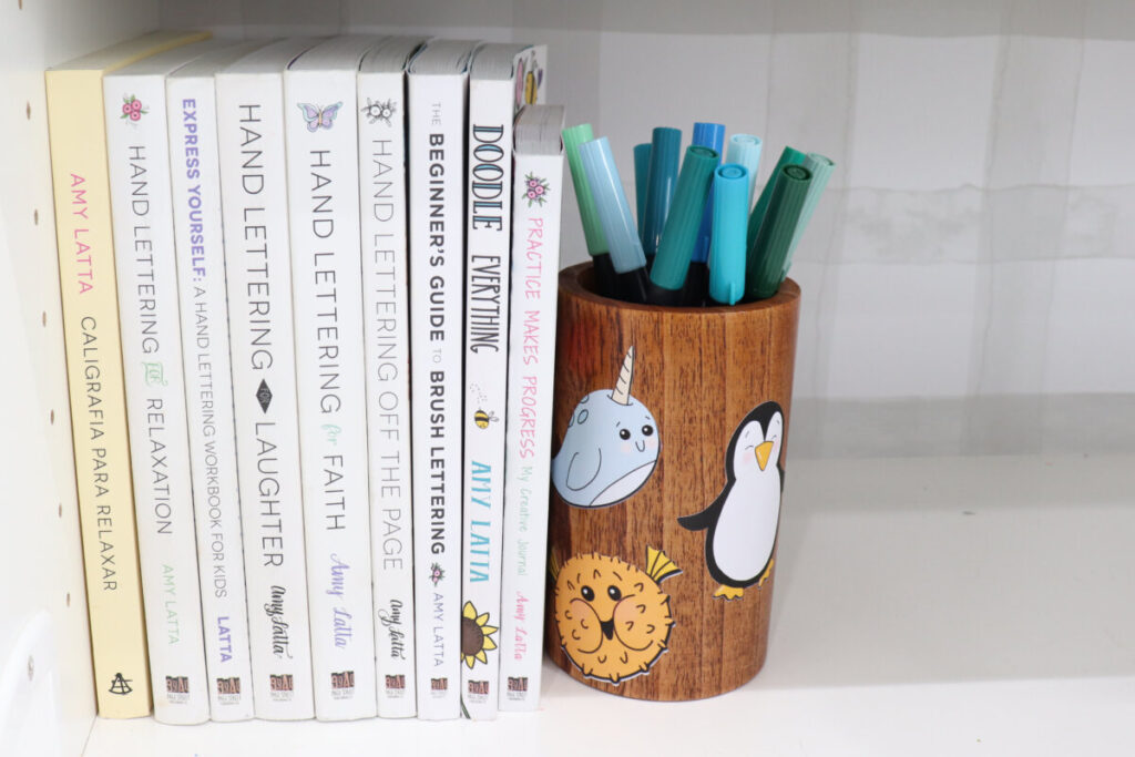 Image contains a vertical stack of nine books, all written by Amy, held up by a wooden pencil holder filled with blue and green markers. The container is covered with animal stickers; a narwhal, a puffer fish, and a penguin.