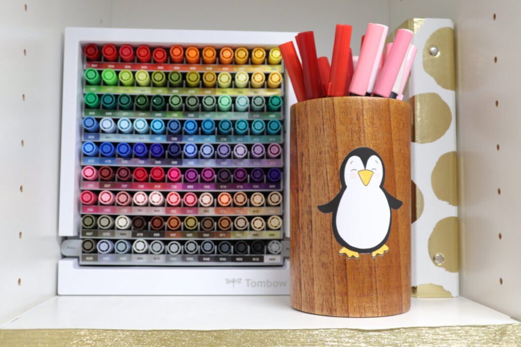 Image contains a marker organizer and a gold and white binder sitting on a white shelf. A wooden pencil holder sits in front, decorated with a penguin sticker, and has pink and red markers inside.