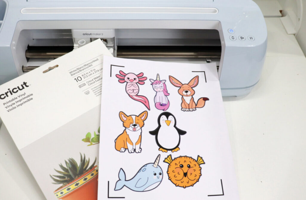 Image contains a blue Cricut Maker 3 sitting on a white table with a pack of Printable Vinyl and a sheet of printed animal stickers on top.