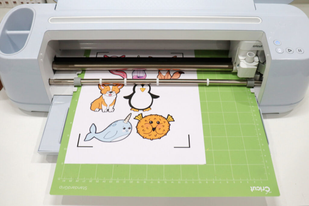 Image contains a blue Cricut Maker loaded with a green cutting mat and a sheet of printed animal stickers in a variety of colors.
