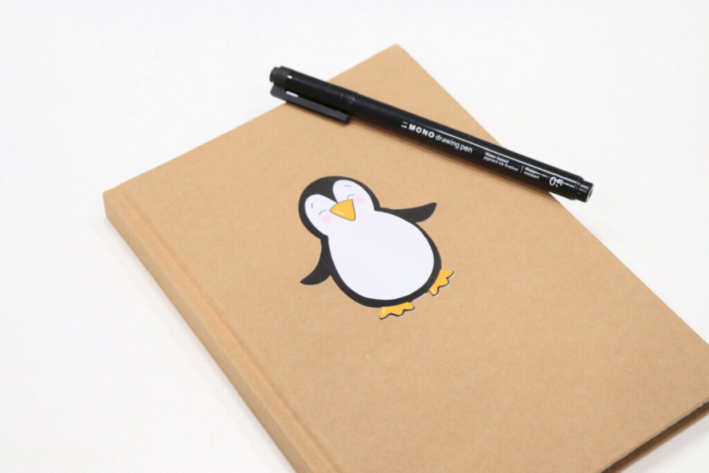 Image contains a kraft colored journal with a penguin sticker applied to the front. It sits on a white table with a black drawing pen on top.