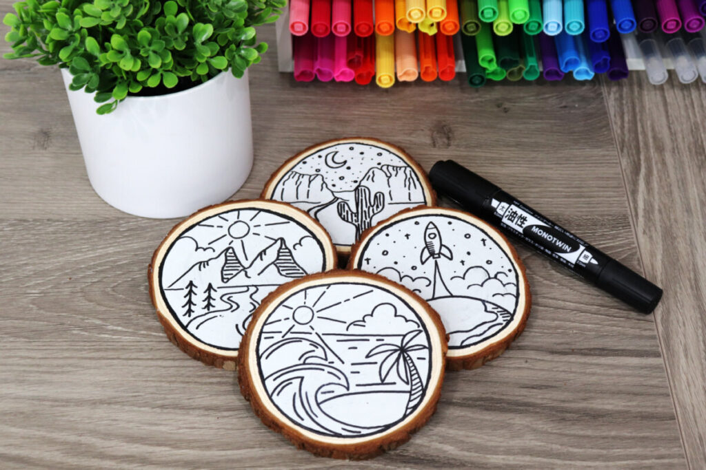 Image contains four wooden coasters, each decorated with a landscape scene drawn in black marker. They sit on a wooden desk with a plant, a black marker, and an organizer filled with colored markers nearby.