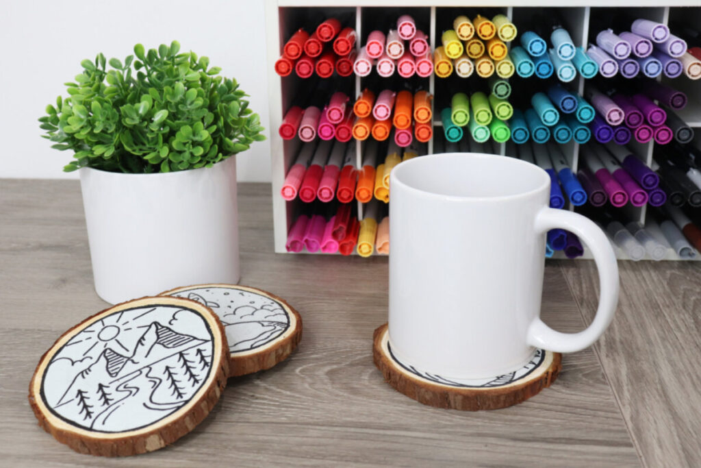Image contains a white coffee mug sitting on a coaster, with two travel coasters and a faux plant nearby. They sit on a wooden desktop with an organizer full of colored markers in the background.