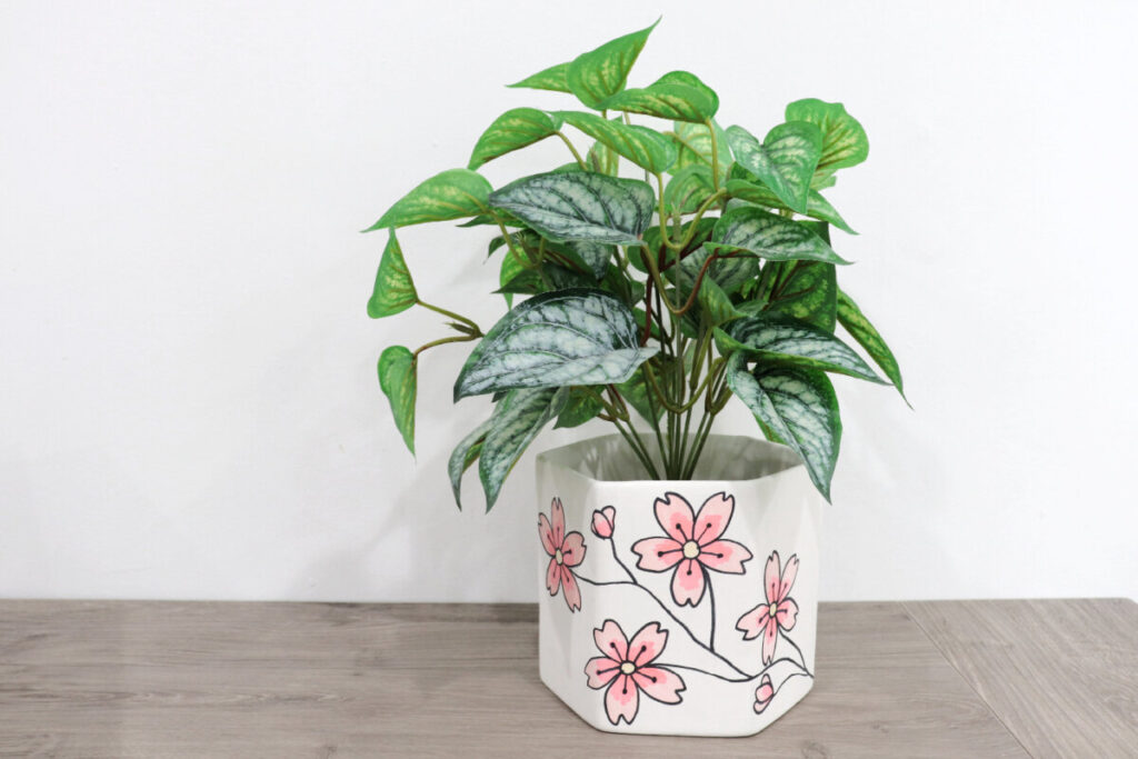 Image contains a faux leafy plant in a hexagon shaped white planter decorated with hand-drawn cherry blossoms.