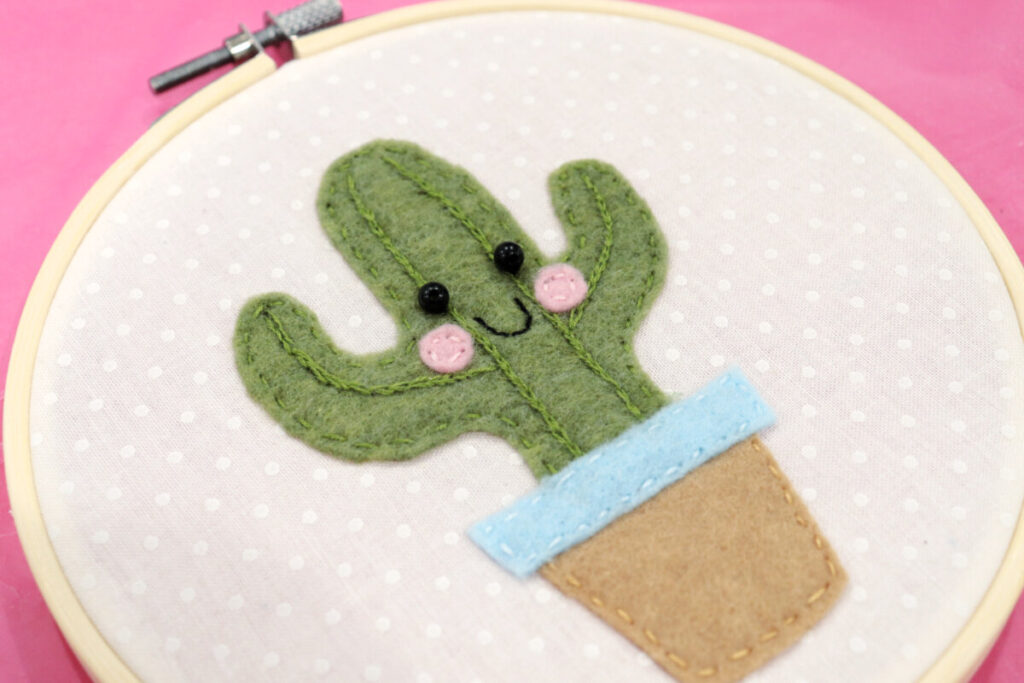 Image contains an embroidery hoop sitting on a pink tabletop with a piece of white fabric inside. In the center of the fabric is a potted felt cactus made from green, brown, blue, and pink felt and black bead eyes.