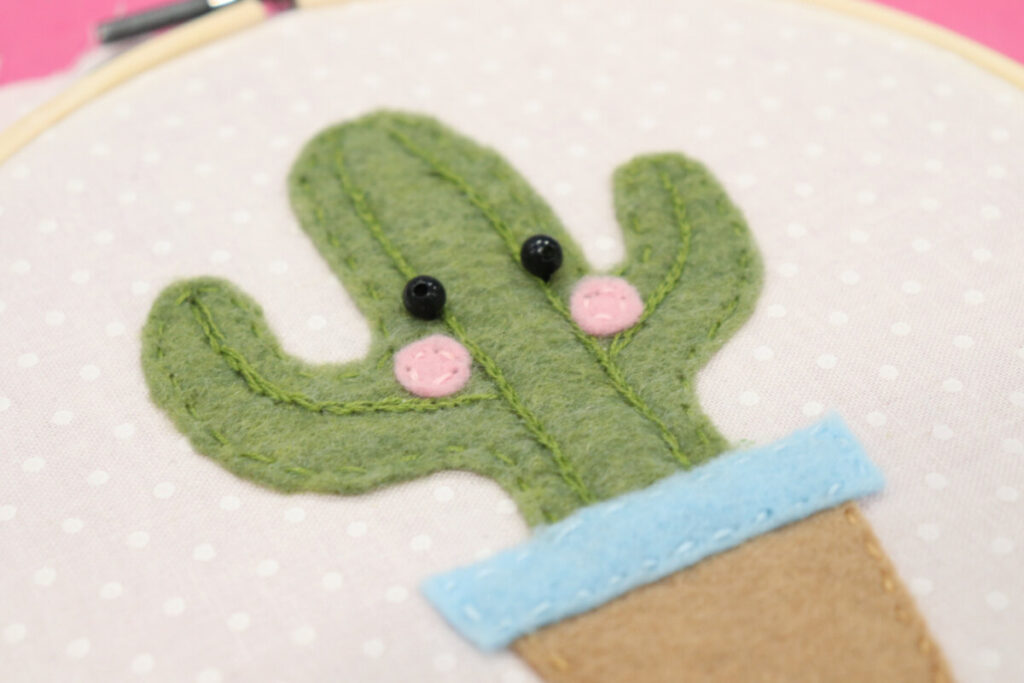 Image contains a green felt cactus in a brown and blue flowerpot stitched onto white fabric. Two black bead eyes are sewn on, as well as two pink felt cheeks.