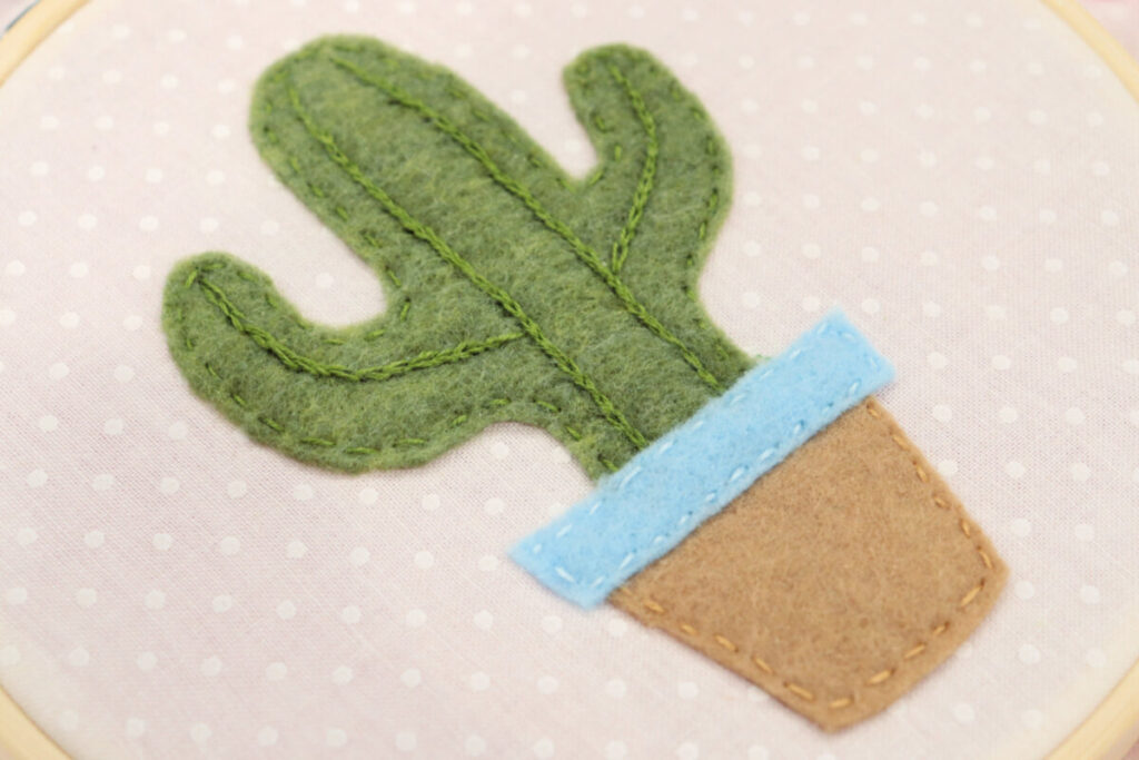 Image contains a closeup of a green felt cactus in a brown and blue felt flowerpot stitched onto a piece of white fabric.