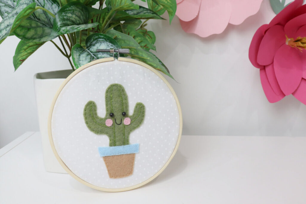 Image contains the finished felt cactus project propped against a faux plant in front of a white wall decorated with large pink paper flowers.