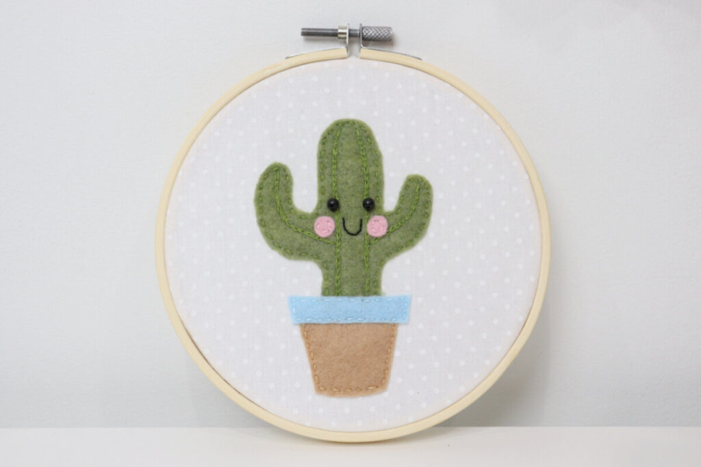 Image contains the finished embroidered felt cactus project propped against a white wall.