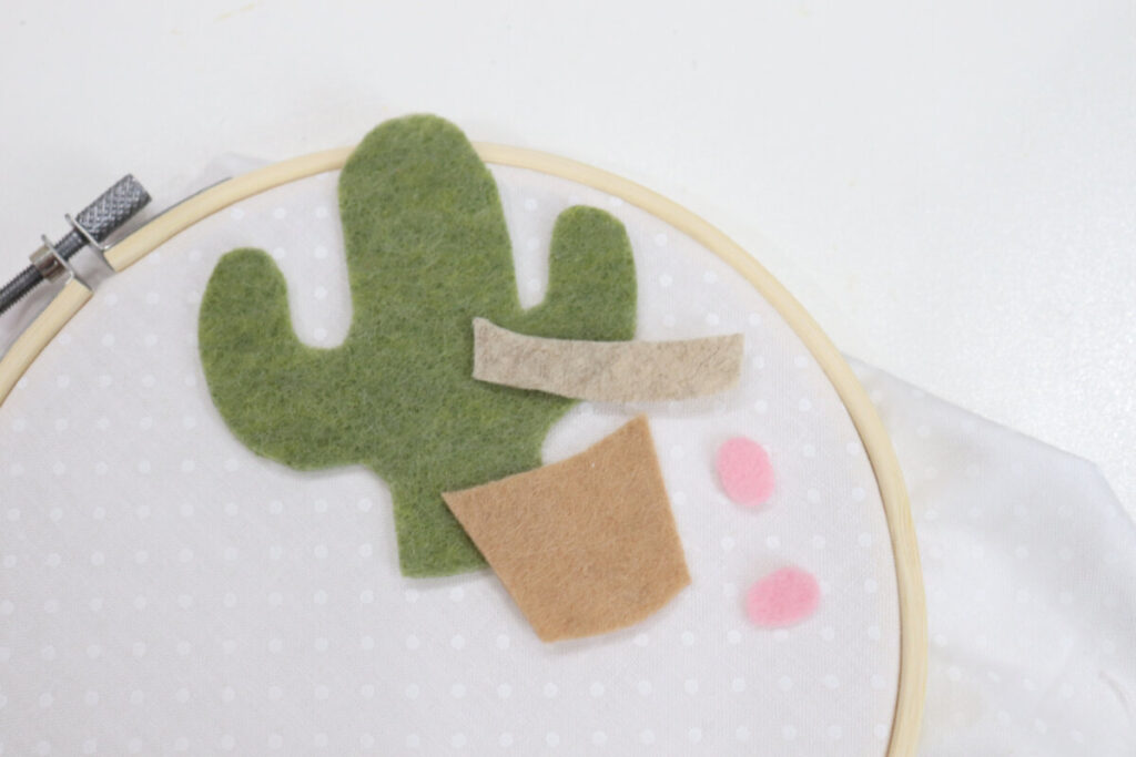 Image contains a bamboo embroidery hoop with white fabric inside, sitting on a white table. Pieces of cut felt sit on top; a green cactus, a brown pot and rim, and two pink ovals.