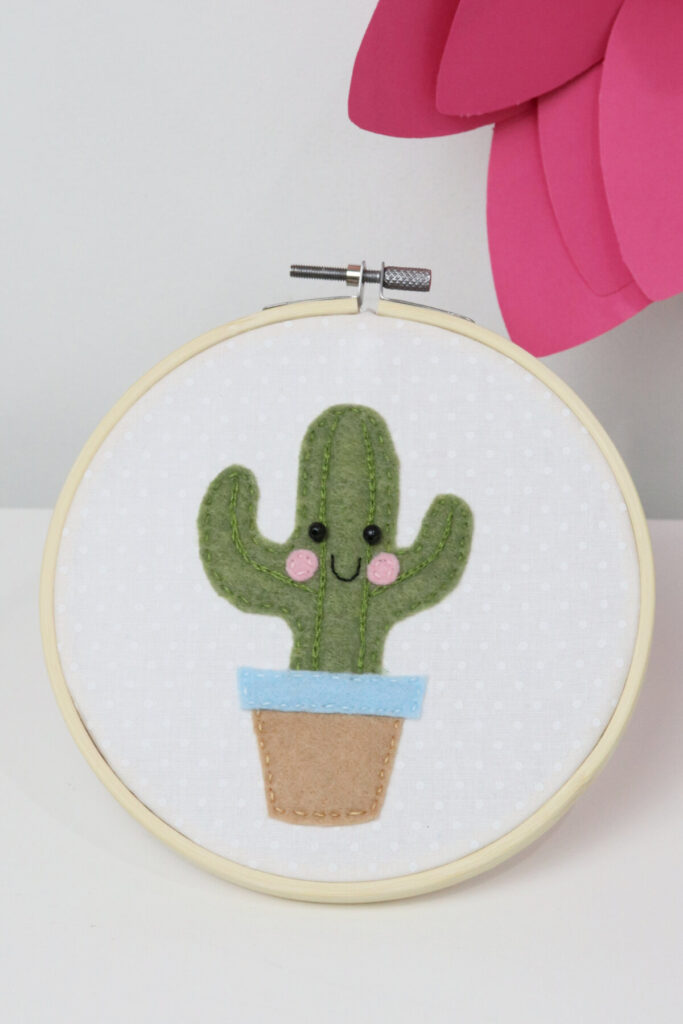 Image contains a wooden embroidery hoop with a piece of white fabric inside. In the center is an embroidered felt cactus with a smiling face.