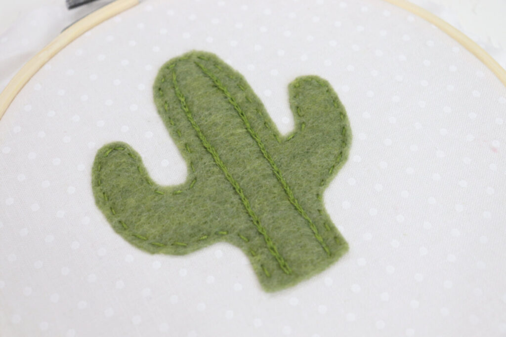 Image is a closeup of the green felt cactus stitched onto a white fabric background. Two texture lines made from reverse chain stitches run through the center of the cactus.