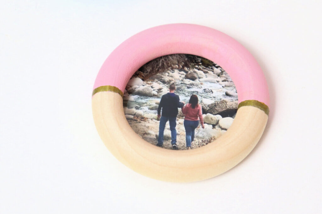 Image contains a wooden circle photo magnet that has been painted pink and gold. A photo of a man and woman walking near a river is inside.
