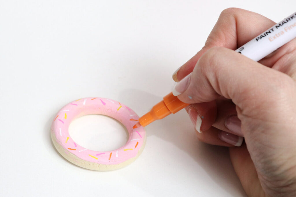 Image contains Amy’s hand holding an orange paint marker and adding sprinkles to a wooden ring painted to look like a pink frosted donut.