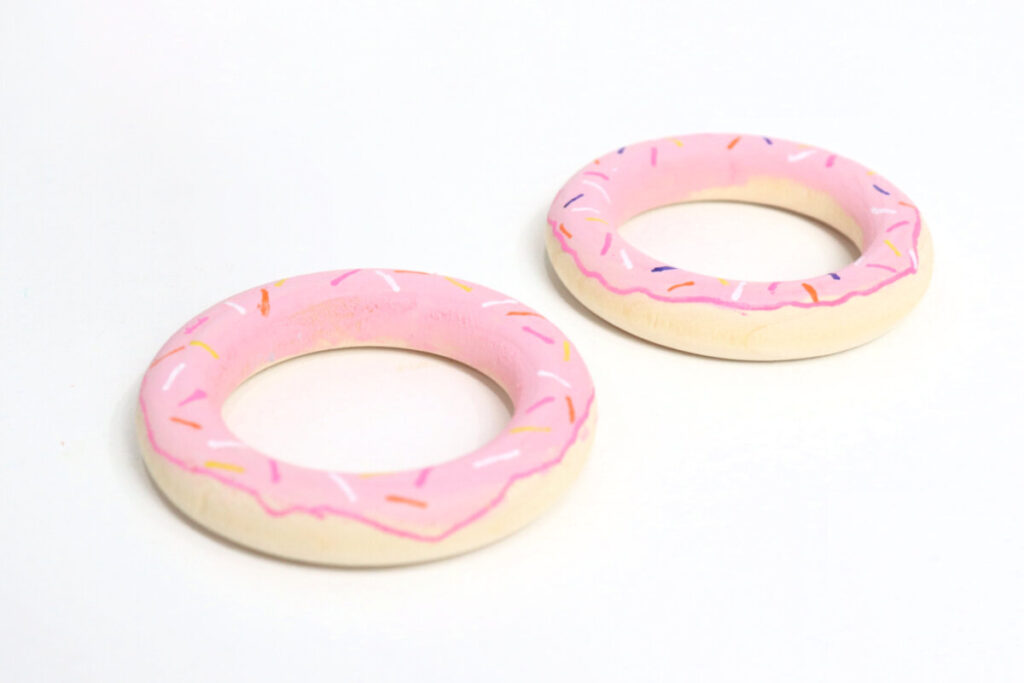 Image contains two wooden rings painted to look like pink frosted donuts with sprinkles.