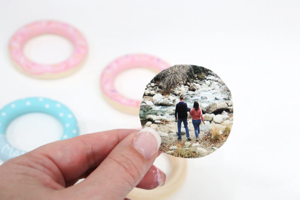 Image contains Amy’s hand holding a photo of a man and woman walking near a river. The photo has been cut into a small circle shape. Painted wooden rings sit in the background on a white table.
