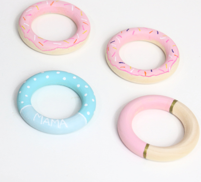 Image contains four wooden rings; two painted to look like pink frosted donuts, one painted half pink with a gold stripe, and one painted teal with white polka dots and the word “mama."