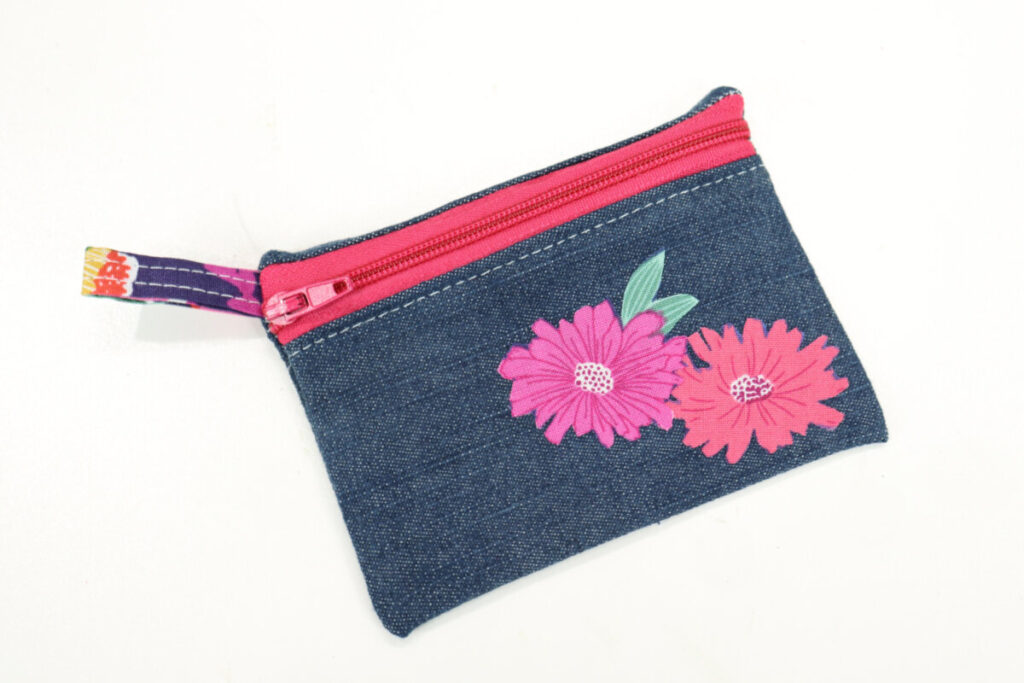 Image contains a small denim zipper pouch with a hot pink zipper and two flowers appliqued to the front.