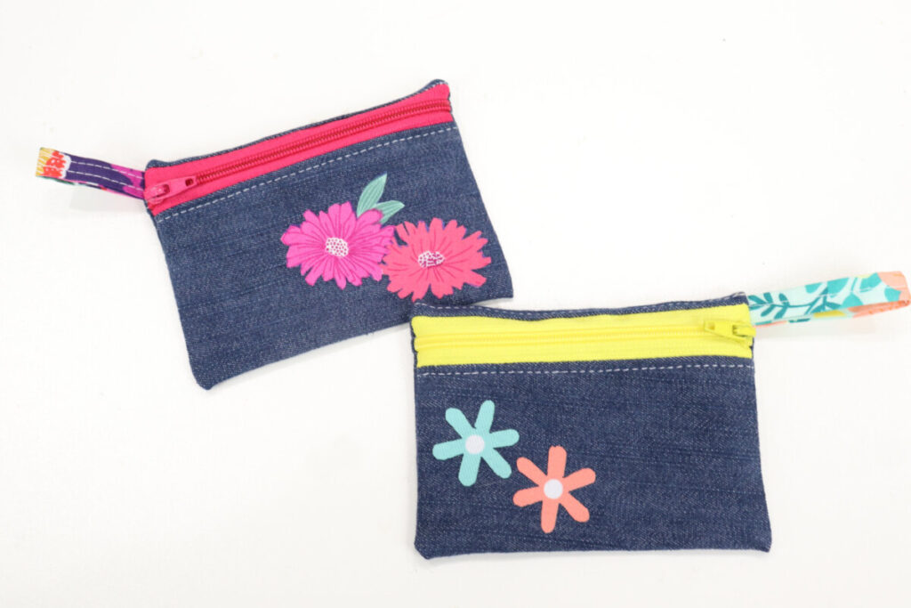 Image contains two finished denim coin purses; one with a pink zipper and pink floral appliques, and one with a yellow zipper and teal and coral flowers.

