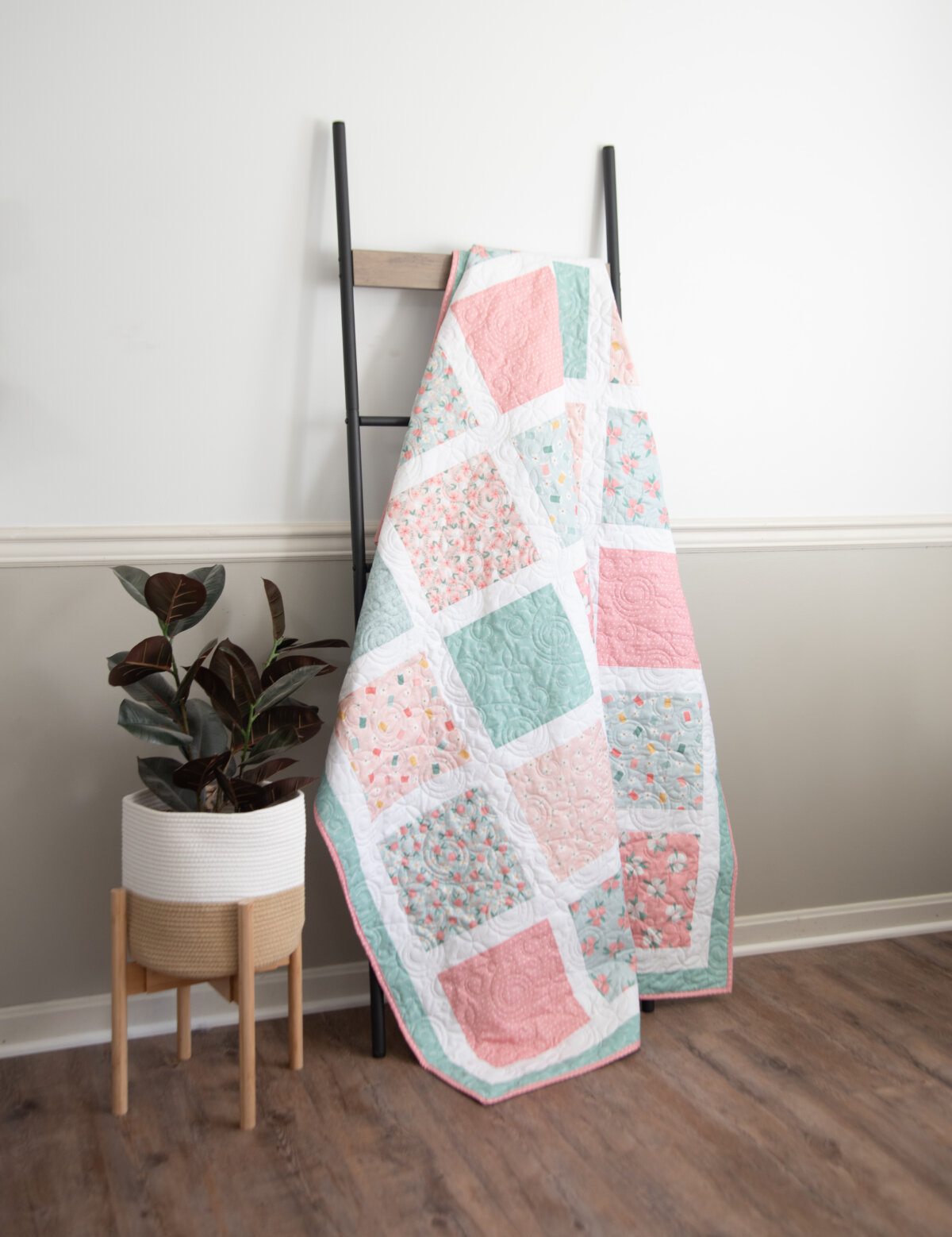 Image contains a large quilt made of pink and green squares on a white background, hanging on a quilt ladder with a faux plant beside it in a stand.