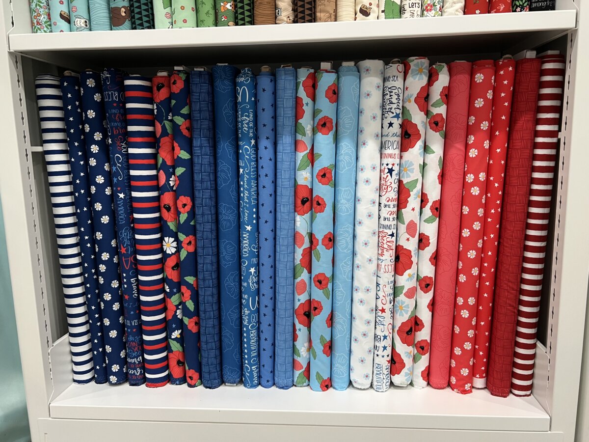 Image contains 24 bolts of assorted patriotic fabric prints from the Oh Say Can You Sew collection by Amy Latta for Andover Fabrics.