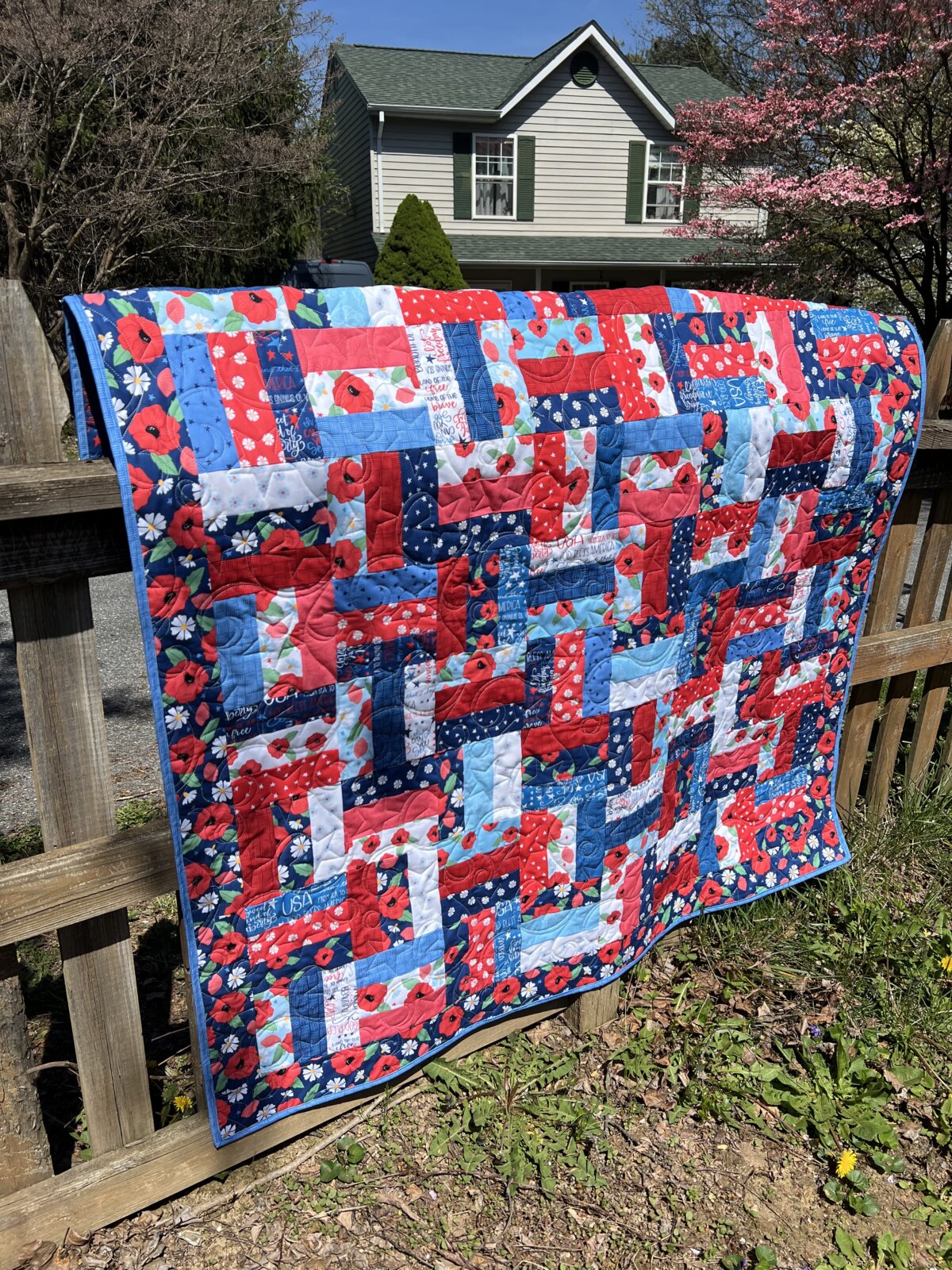 Image contains a red, white, and blue striped quilt on a rail fence in front of a house.