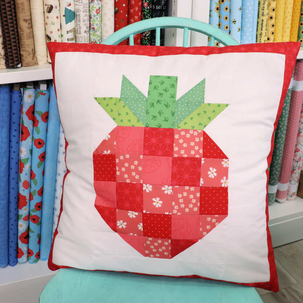 Image contains a white pillow with a red and green patchwork strawberry in the center and a red border.