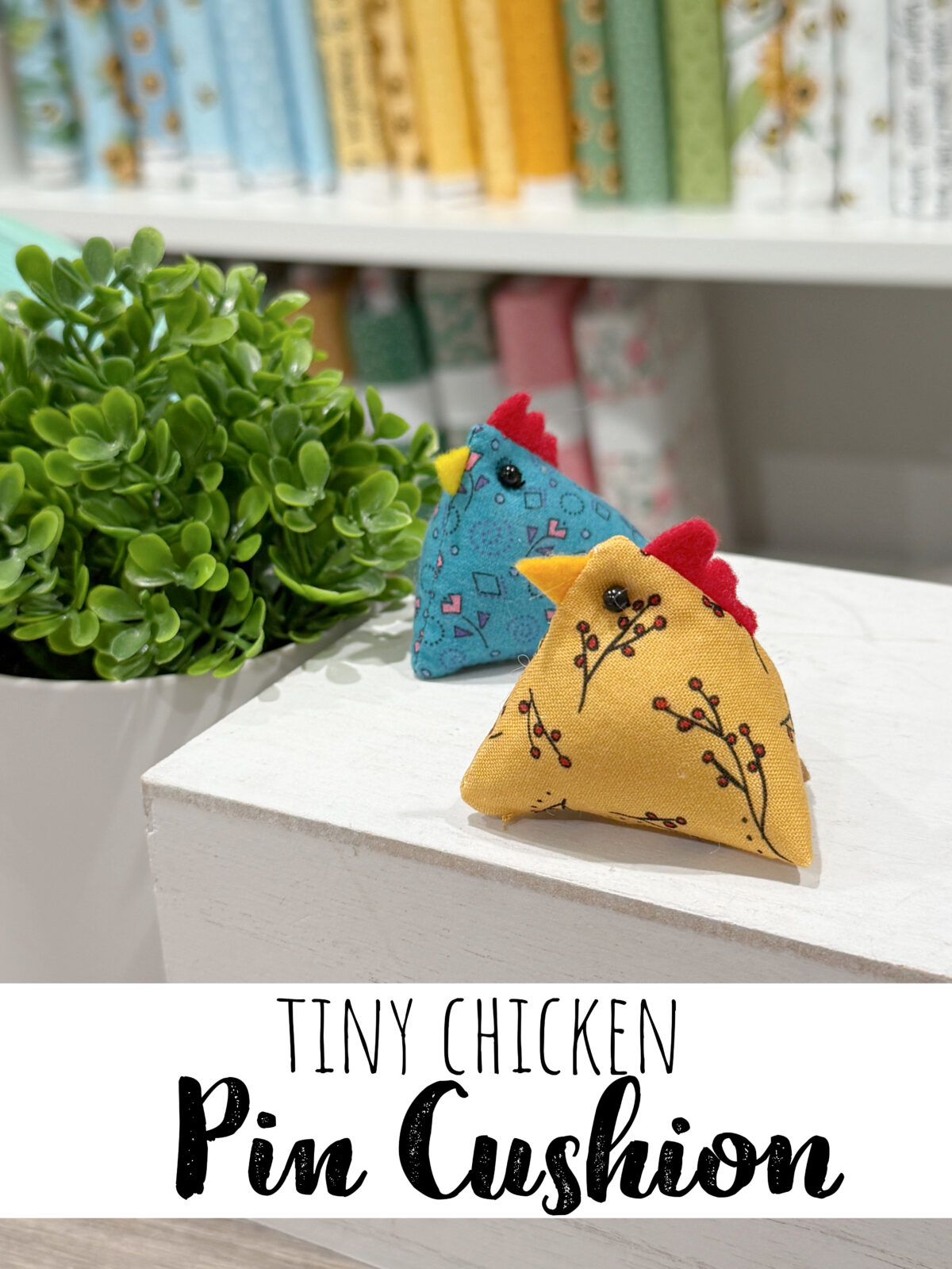 Image contains two tiny chicken pin cushions (one blue, one yellow) sitting on a white box with a faux plant in the background.