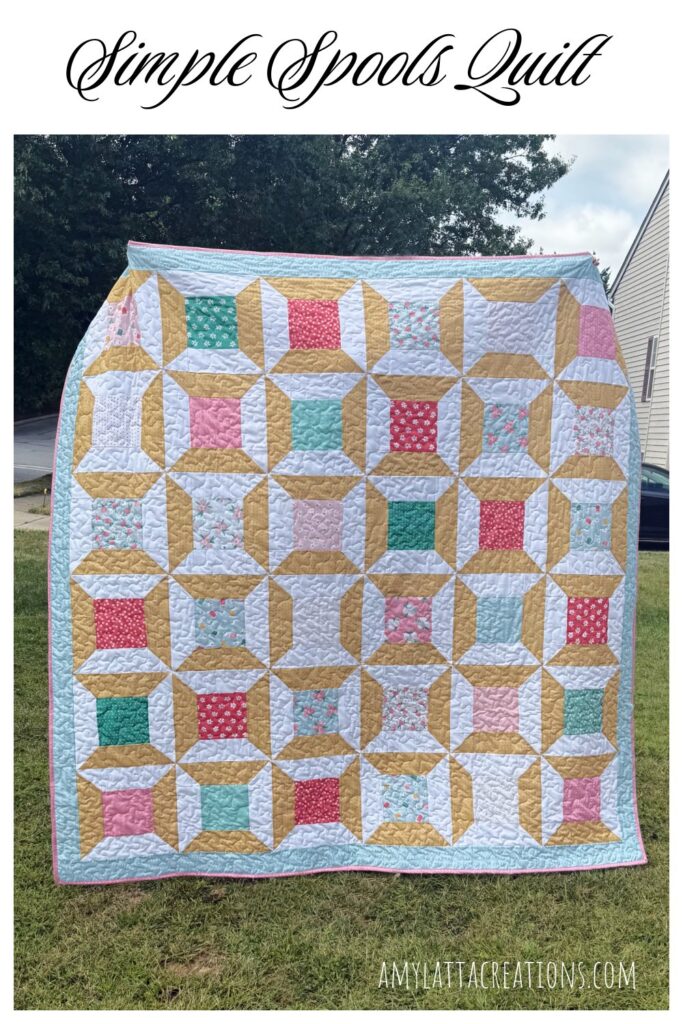 Image contains a quilt made from pink, red, green, and blue fabrics that are designed to look like thread spools.