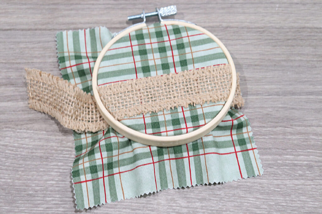 Image contains a miniature wooden hoop with green plaid fabric and burlap ribbon inside, sitting on a wooden desktop.