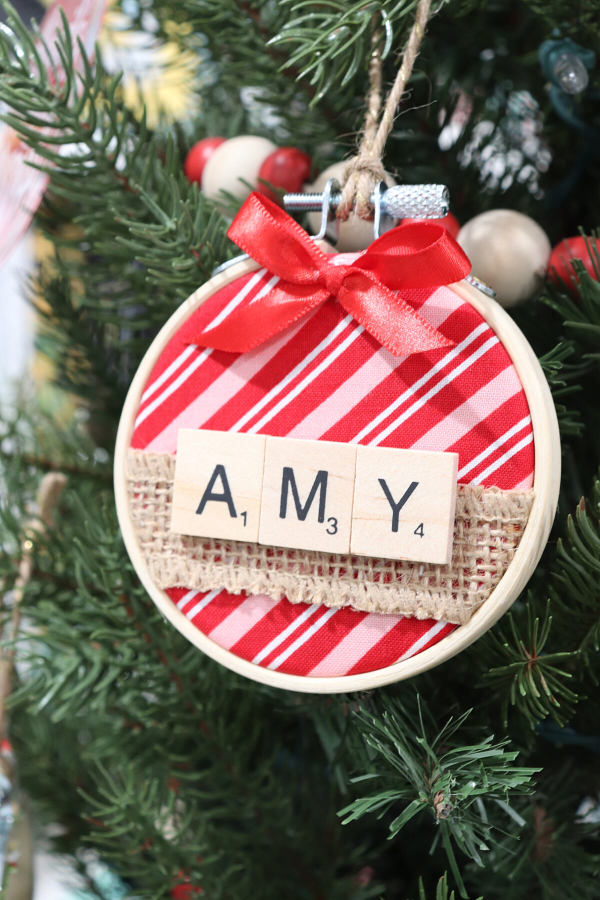Image contains a mini hoop ornament with red, pink, and white striped fabric and the name “Amy” spelled with letter tiles.
