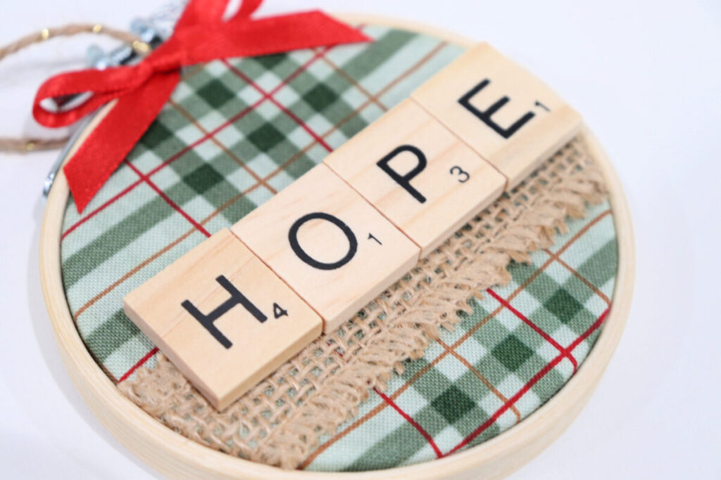 Image contains an ornament made from a wooden hoop, green, plaid fabric, burlap ribbon, and a red bow. Wooden tile letters spell out “hope” across the center.