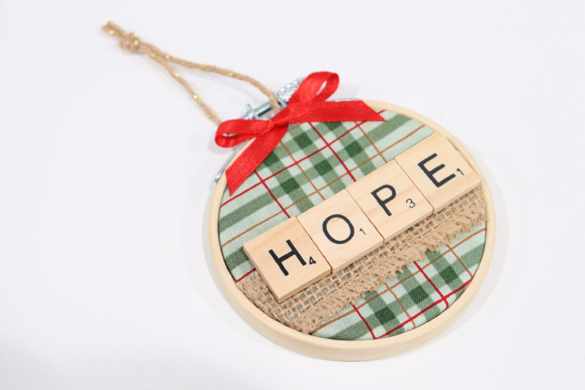 Image contains a mini embroidery hoop with green plaid fabric, burlap ribbon, and wooden letter tiles spelling “hope” inside. A red bow is glued to the top, and it has a twine hanger.