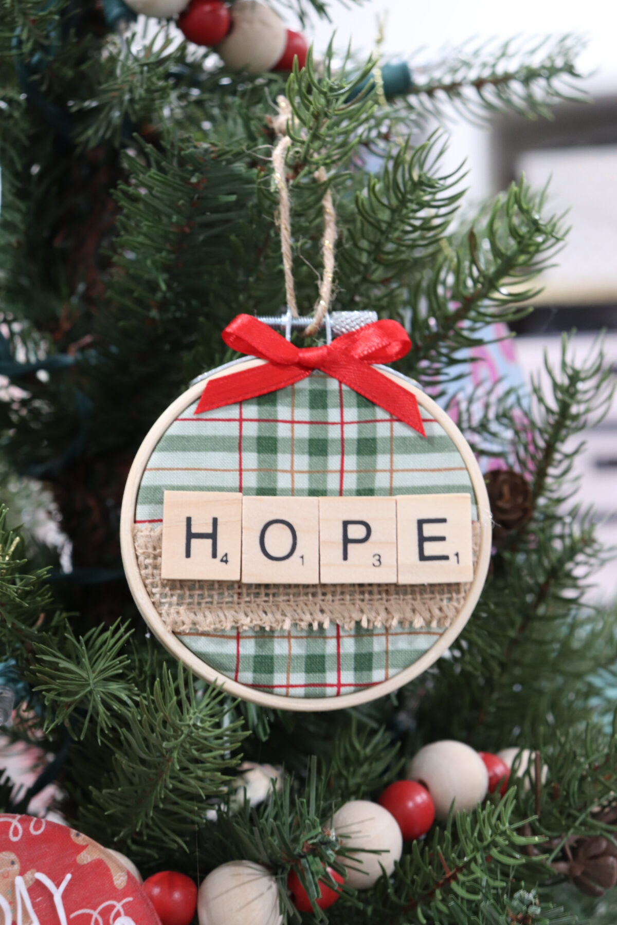 Image contains a mini embroidery hoop containing green plaid fabric, burlap ribbon, and letter tiles spelling out “hope.” A red bow is at the top of the ornament, along with a twine hanger. The ornament hangs on an artificial tree.